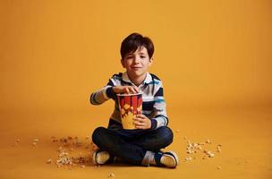 Cute little boy sitting and watching movie and eats popcorn against yellow wall photo