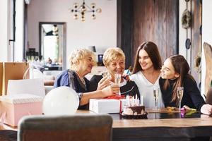 Glasses with alcohol in hands and cake on table. Senior woman with family and friends celebrating a birthday indoors photo