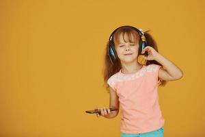 Portrait of cute little girl in headphones and with phone in the studio against yellow background photo