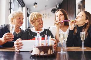 Glasses with alcohol in hands and cake on table. Senior woman with family and friends celebrating a birthday indoors photo