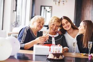 Glasses with alcohol in hands and cake on table. Senior woman with family and friends celebrating a birthday indoors photo