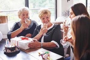 vasos con alcohol en las manos y pastel en la mesa. mujer mayor con familiares y amigos celebrando un cumpleaños en el interior foto