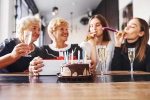 Glasses with alcohol in hands and cake on table. Senior woman with family and friends celebrating a birthday indoors photo