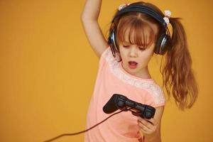 Little female gamer in headphones and with joystick in hands playing video games against yellow background photo