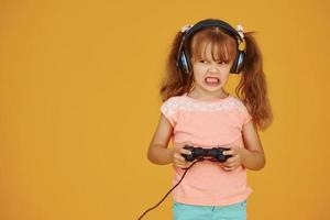 Little female gamer in headphones and with joystick in hands playing video games against yellow background photo