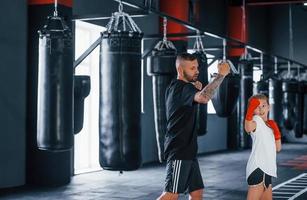 golpes básicos. joven entrenador de boxeo tatuado enseña a una linda niñita en el gimnasio foto