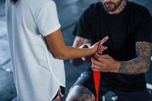 ayudando a llevar las vendas. joven entrenador de boxeo tatuado enseña a una linda niñita en el gimnasio foto