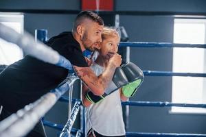 Standing on the boxing ring. Young tattooed boxing coach teaches cute little girl in the gym photo