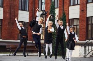 grupo de niños en uniforme escolar saltando y divirtiéndose juntos al aire libre cerca del edificio educativo foto