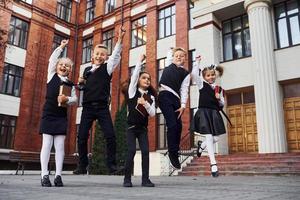 Group of kids in school uniform jumping and having fun outdoors together near education building photo