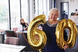 con globos del número 60 en las manos. mujer mayor con familiares y amigos celebrando un cumpleaños en el interior foto