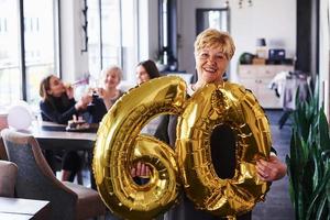 With balloons of number 60 in hands. Senior woman with family and friends celebrating a birthday indoors photo