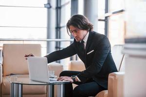 Sits on the sofa with laptop. Portrait of handsome young businessman in black suit and tie photo