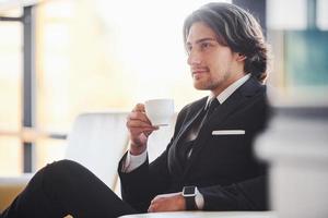 se sienta en el sofá con una taza de bebida. retrato de un apuesto joven hombre de negocios con traje negro y corbata