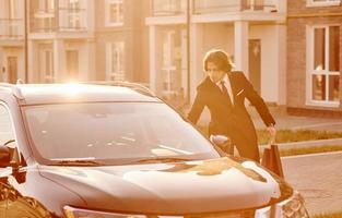 Portrait of handsome young businessman in black suit and tie outdoors near modern car in the city photo