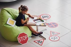 una linda niña se sienta en la suave silla verde junto a la mesa con lápiz y hojas de papel. cerca de automóviles modernos y señales de tráfico en el suelo foto