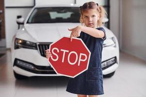 Portrait of cute little girl that holds road sign in hands in automobile salon photo