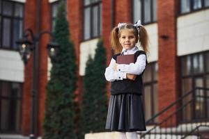 Young schoolgirl in formal wear and notepad in hands stands near the building outdoors photo