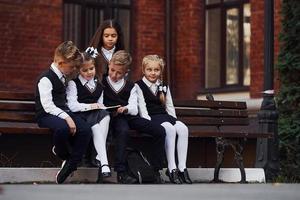 niños de la escuela en uniforme que se sienta al aire libre en el banco con bloc de notas foto