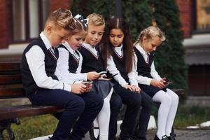 niños de la escuela en uniforme que se sienta al aire libre en el banco con teléfonos inteligentes foto