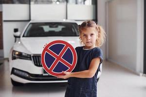 Portrait of cute little girl that holds road sign in hands in automobile salon photo