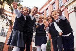 divirtiéndose y abrazándose unos a otros. grupo de niños con uniforme escolar que están juntos al aire libre cerca del edificio de educación foto