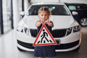 Portrait of cute little girl that holds road sign in hands in automobile salon photo