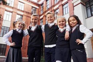 divirtiéndose y abrazándose unos a otros. grupo de niños con uniforme escolar que están juntos al aire libre cerca del edificio de educación foto