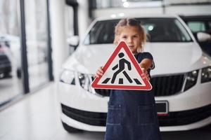 Portrait of cute little girl that holds road sign in hands in automobile salon photo