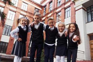 divirtiéndose y abrazándose unos a otros. grupo de niños con uniforme escolar que están juntos al aire libre cerca del edificio de educación foto