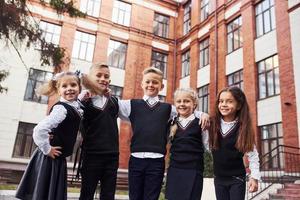 divirtiéndose y abrazándose unos a otros. grupo de niños con uniforme escolar que están juntos al aire libre cerca del edificio de educación foto