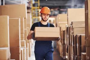 joven trabajador de almacenamiento en uniforme y casco lleva caja en las manos foto