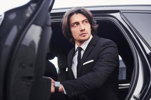 Portrait of handsome young businessman in black suit and tie walks out from modern car photo