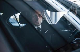 joven hombre de negocios con traje negro y corbata conduciendo un automóvil moderno foto