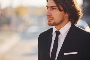 Portrait of handsome young businessman in black suit and tie stands in the city at sunny day photo