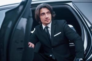 Portrait of handsome young businessman in black suit and tie walks out from modern car photo