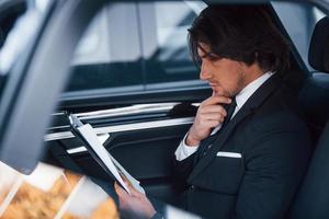 Using notepad. Young businessman in black suit and tie inside modern automobile photo