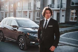 Portrait of handsome young businessman in black suit and tie outdoors near modern car in the city photo
