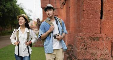 feliz pareja de viajeros asiáticos con sombrero y mochila visitan el templo antiguo. sonriente joven y mujer caminando con cámara. concepto de vacaciones, viajes, relajación y hobby. video