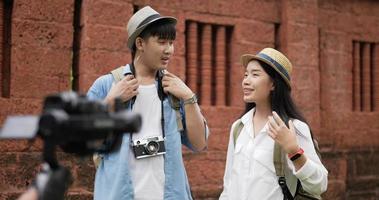 Funny asian traveler couple with hat talking and looking at camera at ancient temple. Smiling young man and woman videoblogger filming new vlog video with professional camera at ancient temple.