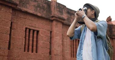 Side view of Happy asian traveler man with hat takes a photo and visits ancient temple. Smiling young male walking and holding camera. Holiday, travel and hobby concept. video