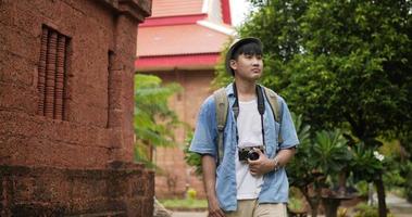 Happy asian traveler man with hat visiting ancient temple. Smiling young male walking and holding camera. Holiday, travel and hobby concept. video