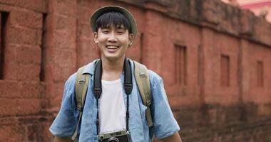 Headshot of Asian traveler man with hat talking and looking at camera at ancient temple. Smiling male videoblogger filming new vlog video with professional camera at ancient temple. Travel and hobby.