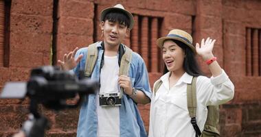 Funny asian traveler couple with hat talking and looking at camera at ancient temple. Smiling young man and woman videoblogger filming new vlog video with professional camera at ancient temple.