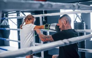 Having sparring with each other on the boxing ring. Young tattooed boxing coach teaches cute little girl in the gym photo