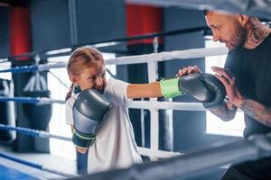 tener sparring entre sí en el ring de boxeo. joven entrenador de boxeo tatuado enseña a una linda niñita en el gimnasio foto