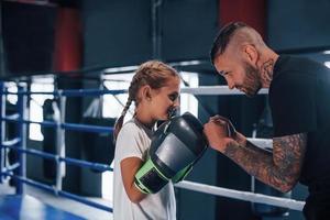 tener sparring entre sí en el ring de boxeo. joven entrenador de boxeo tatuado enseña a una linda niñita en el gimnasio foto