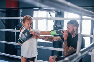 Having sparring with each other on the boxing ring. Young tattooed boxing coach teaches cute little girl in the gym photo