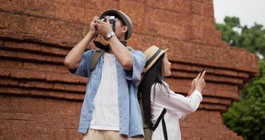 Happy asian traveler couple with hat takes a photo and visits ancient temple. Smiling young man and woman standing and looking ancient temple. Holiday, travel and hobby concept. video
