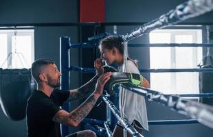 concepción de la asistencia. joven entrenador de boxeo tatuado enseña a una linda niñita en el gimnasio foto